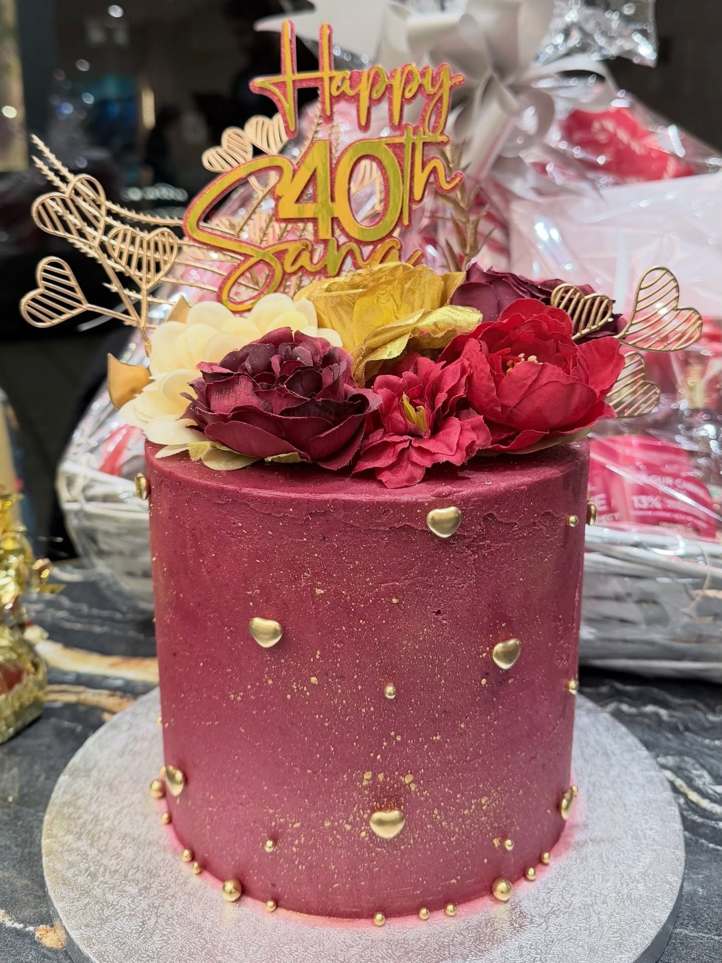 Deep burgundy 40th birthday cake decorated with gold hearts, rich red florals and a gold topper