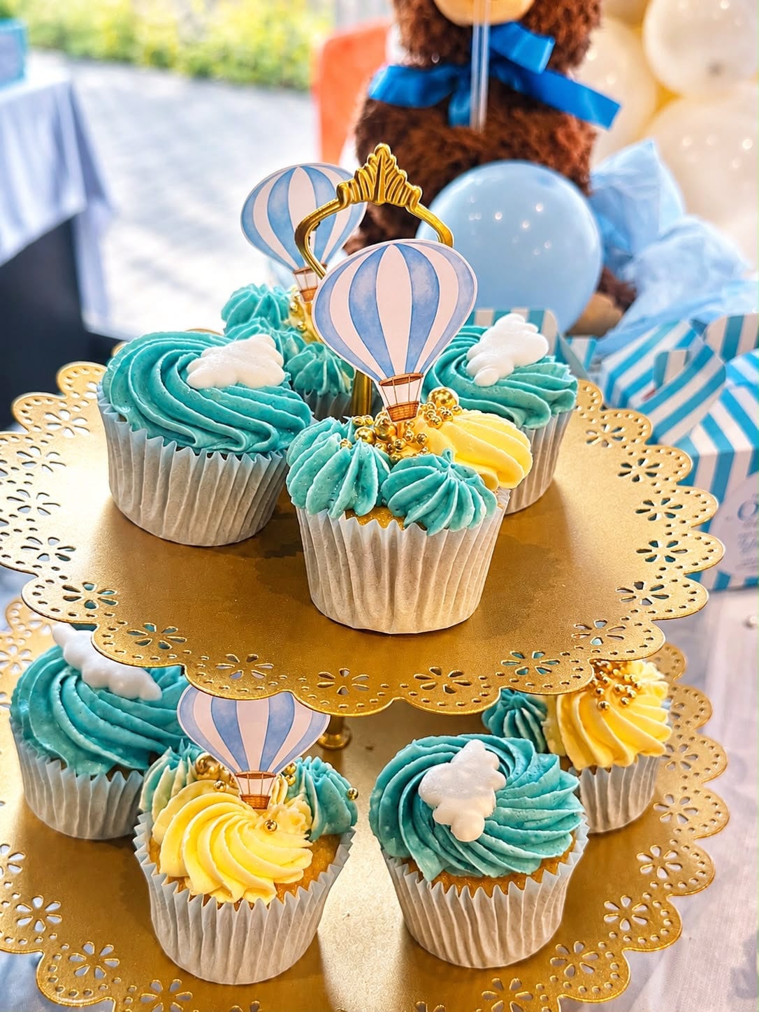 Teal and yellow buttercream cupcakes with hot air balloon and cloud toppers on a gold stand