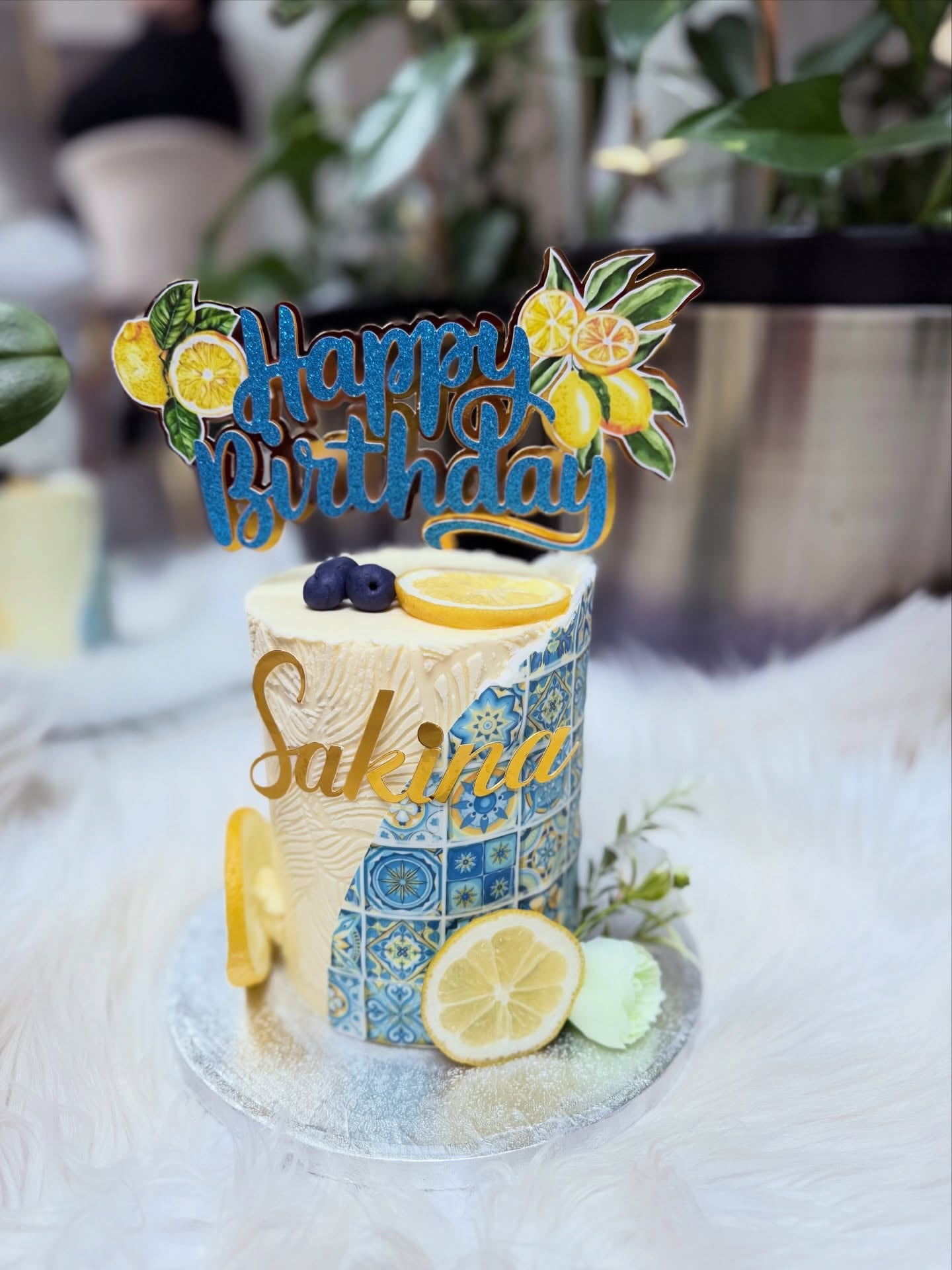 Ivory buttercream birthday cake with a blue tile panel, fresh lemon slices, blueberries and a gold topper