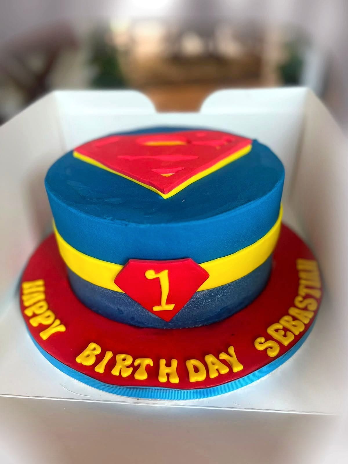 Superhero Emblem First Birthday Cake by SpongyBakes — A bold superhero-emblem cake with a red fondant band and piped lettering.