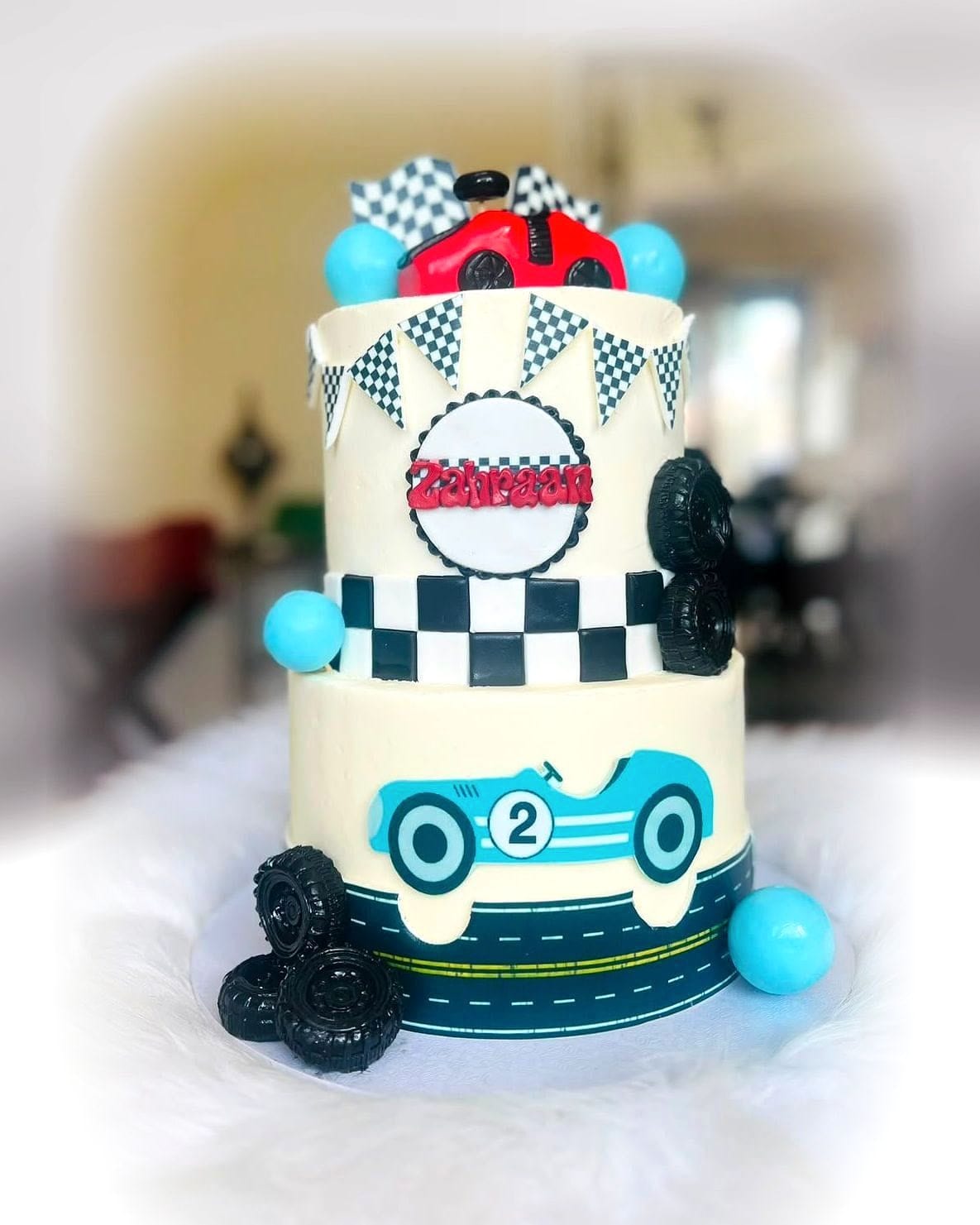 Race Day Two-Tier Cake by SpongyBakes — A two-tier racing cake with a chequered flag band, fondant tyres and a car topper.