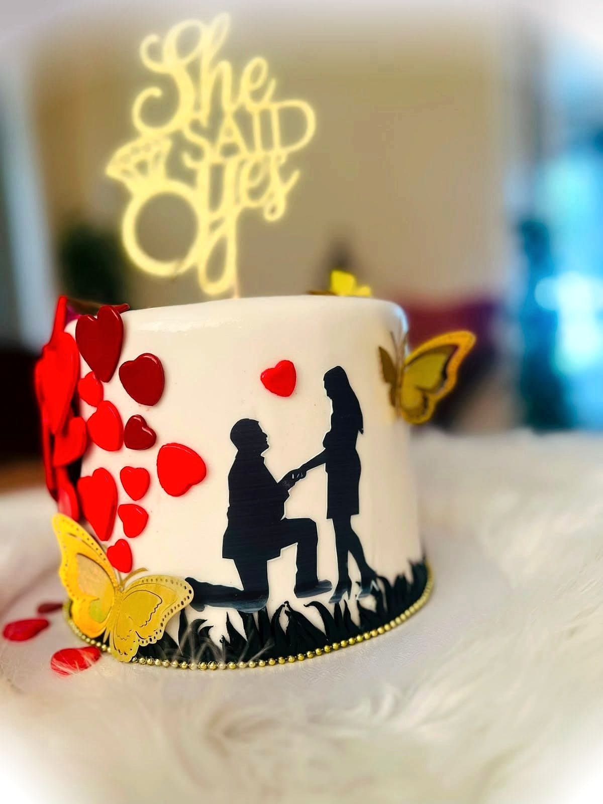 She Said Yes Engagement Cake by SpongyBakes — An ivory cake with a silhouette proposal scene, fondant hearts and a gold topper.