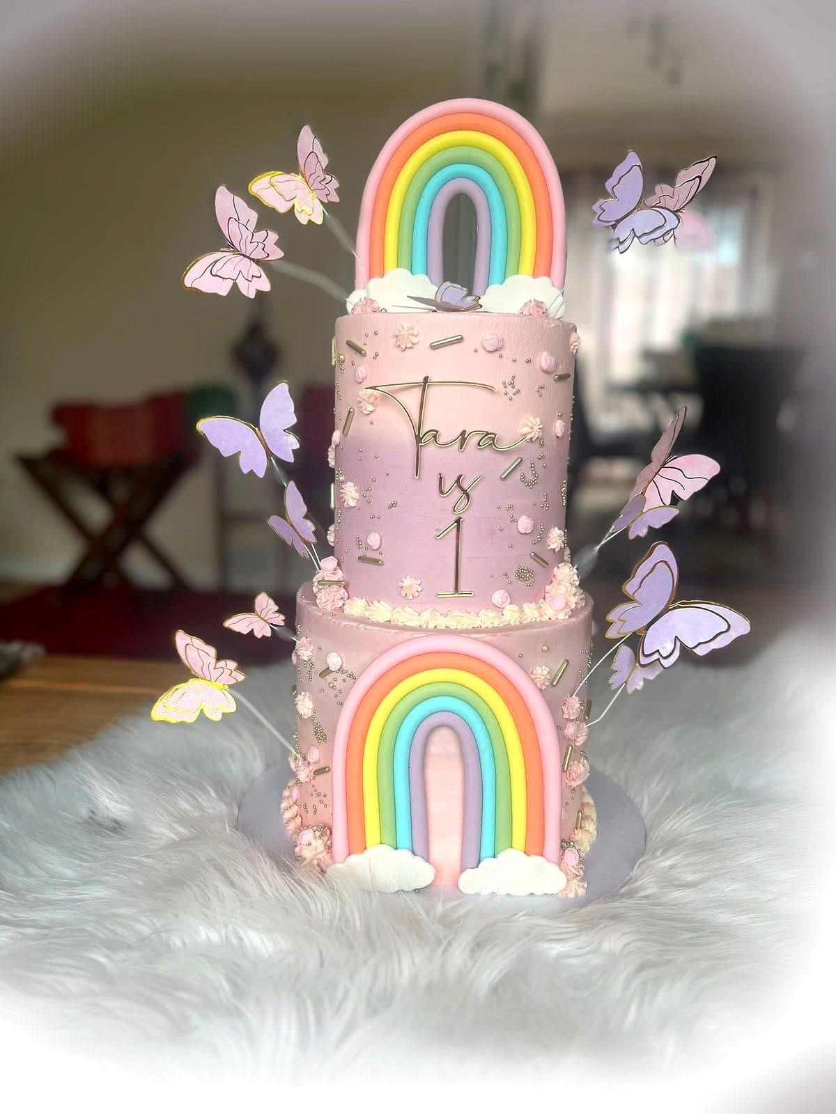 Rainbow Butterfly First Birthday Cake by SpongyBakes — A two-tier pink cake with a fondant rainbow, butterflies and a textured finish.